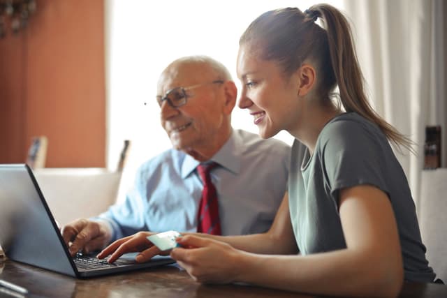 A young woman leans in to help an older man with something on his laptop at home