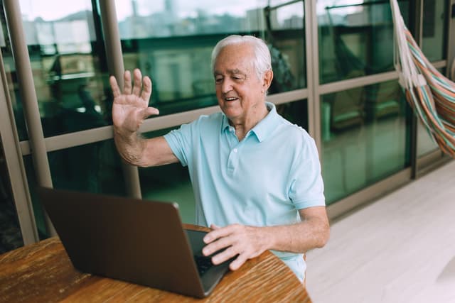 A smiling older man waves hello to someone on a video call on his laptop